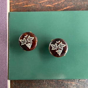May include: Two round, brown, decorative buttons with a gold trim. Each button features a white, etched design of a grapevine with leaves and grapes.