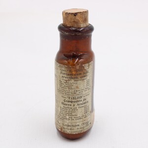 May include: A small, antique brown glass bottle with a cork stopper. The bottle has a label with faded text, including the word "TABLOID" and the phrase "Compuesto de Hierro y Arsenico". The bottle is likely a vintage medicine container.