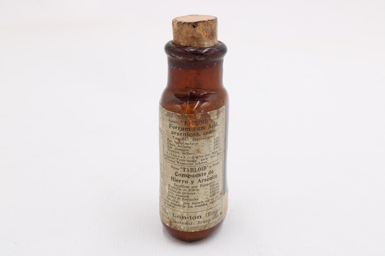May include: A small, antique brown glass bottle with a cork stopper. The bottle has a label with faded text, including the word "TABLOID" and the phrase "Compuesto de Hierro y Arsenico". The bottle is likely a vintage medicine container.