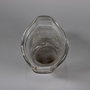 May include: Clear glass pitcher with a wide, curved spout and a rounded body. The pitcher is empty and appears to be clean. The design is simple and elegant, suitable for serving beverages. The pitcher is set against a plain, light gray background.