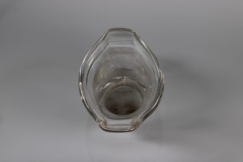 May include: Clear glass pitcher with a wide, curved spout and a rounded body. The pitcher is empty and appears to be clean. The design is simple and elegant, suitable for serving beverages. The pitcher is set against a plain, light gray background.