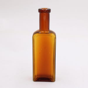 May include: Amber-colored glass bottle with a rectangular body and a long, cylindrical neck. The bottle has a small opening at the top and is made of transparent glass. The bottle is empty and sits on a white surface.