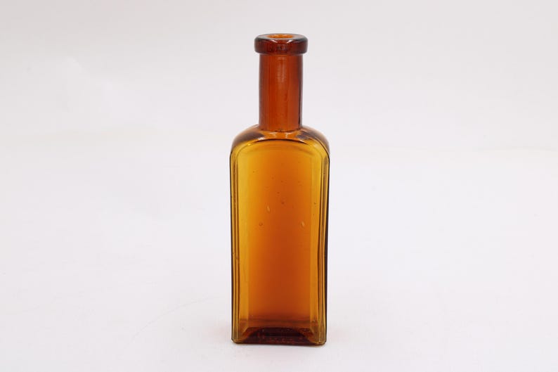 May include: Amber-colored glass bottle with a rectangular body and a long, cylindrical neck. The bottle has a small opening at the top and is made of transparent glass. The bottle is empty and sits on a white surface.