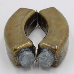 May include: Two antique brass flasks with a curved design, connected by a hinge. The flasks have a tarnished, aged appearance, with a silver-colored cap on each end. The flasks are displayed against a white background.