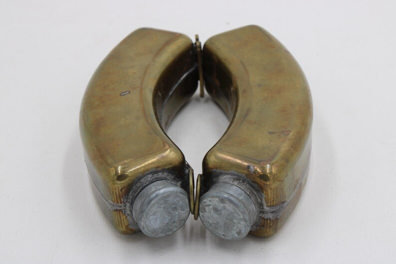May include: Two antique brass flasks with a curved design, connected by a hinge. The flasks have a tarnished, aged appearance, with a silver-colored cap on each end. The flasks are displayed against a white background.
