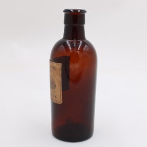 Puede incluir: Una botella de vidrio marr&oacute;n con una etiqueta. La botella tiene un cuerpo redondeado y un cuello estrecho. La etiqueta es rectangular y parece tener texto y un dise&ntilde;o. La botella est&aacute; vac&iacute;a y se encuentra sobre una superficie blanca.