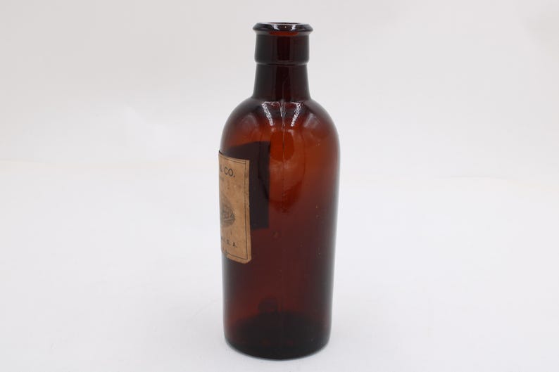 Puede incluir: Una botella de vidrio marr&oacute;n con una etiqueta. La botella tiene un cuerpo redondeado y un cuello estrecho. La etiqueta es rectangular y parece tener texto y un dise&ntilde;o. La botella est&aacute; vac&iacute;a y se encuentra sobre una superficie blanca.