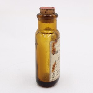 May include: An amber glass bottle with a cork stopper. The bottle has a label with faded text. The bottle is cylindrical with a rounded base and a narrow neck. The cork is reddish-brown.