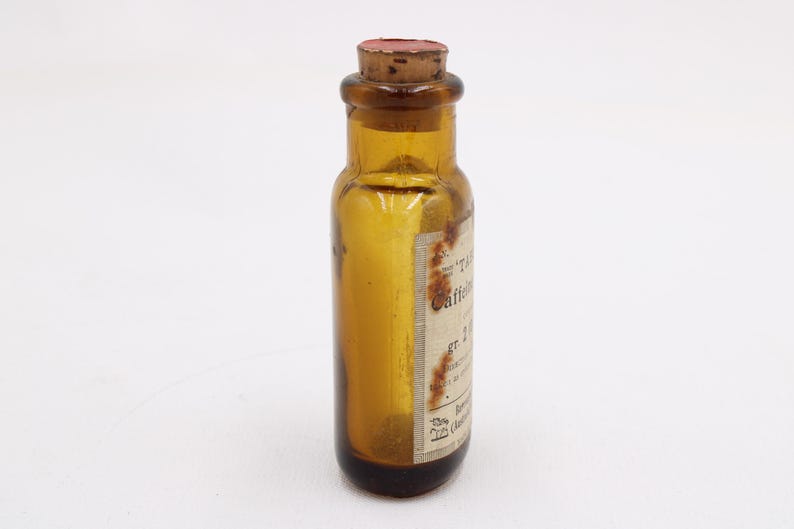 May include: An amber glass bottle with a cork stopper. The bottle has a label with faded text. The bottle is cylindrical with a rounded base and a narrow neck. The cork is reddish-brown.