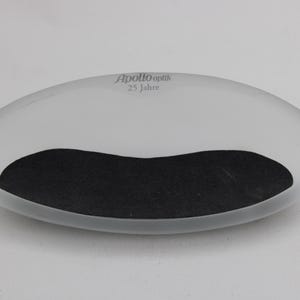K&ouml;nnte beinhalten: Eine ovale, mattierte Schale mit einem schwarzen, gebogenen Einsatz. Der Text "Apollo Optik 25 Jahre" ist grau auf der Oberseite aufgedruckt. Die Schale steht vor einem schlichten wei&szlig;en Hintergrund.