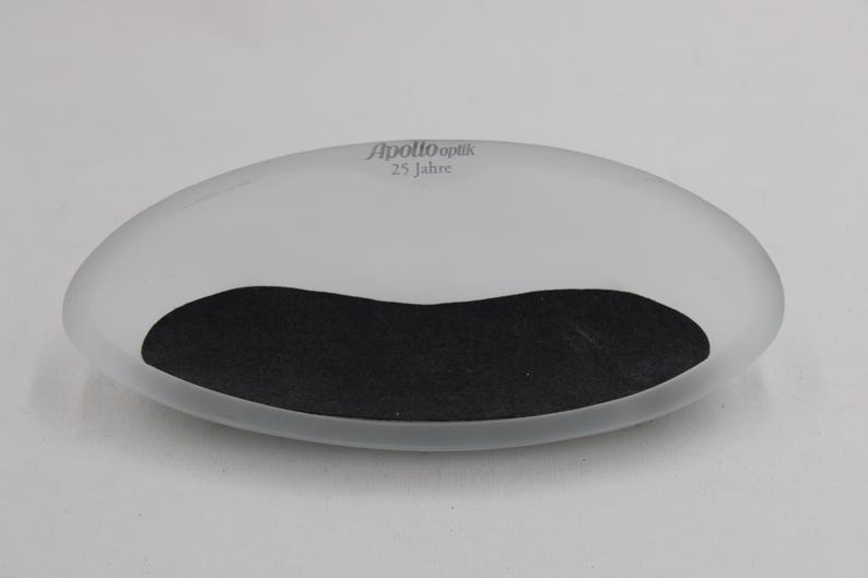 K&ouml;nnte beinhalten: Eine ovale, mattierte Schale mit einem schwarzen, gebogenen Einsatz. Der Text "Apollo Optik 25 Jahre" ist grau auf der Oberseite aufgedruckt. Die Schale steht vor einem schlichten wei&szlig;en Hintergrund.