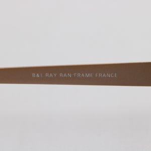 May include: Close-up of a section of a pair of eyeglasses. The frame is a light brown color. The text "B&I RAY BAN FRAME FRANCE" is printed on the frame.