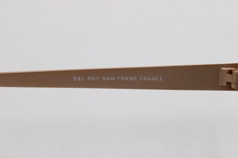 May include: Close-up of a section of a pair of eyeglasses. The frame is a light brown color. The text "B&I RAY BAN FRAME FRANCE" is printed on the frame.