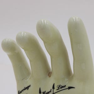 May include: A cream-coloured ceramic hand figurine, featuring black text and arrows indicating palmistry lines. The fingers are slightly curved, and the surface has a glossy finish. The words "Marriage", "Heart Line", and other text are visible.