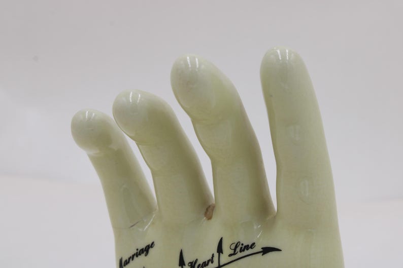 May include: A cream-coloured ceramic hand figurine, featuring black text and arrows indicating palmistry lines. The fingers are slightly curved, and the surface has a glossy finish. The words "Marriage", "Heart Line", and other text are visible.