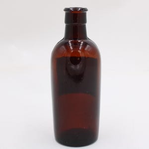 Puede incluir: Una botella de vidrio marr&oacute;n con un cuerpo redondeado y un cuello estrecho. La botella est&aacute; vac&iacute;a y tiene un color &aacute;mbar oscuro. La botella probablemente se utiliza para almacenar l&iacute;quidos.