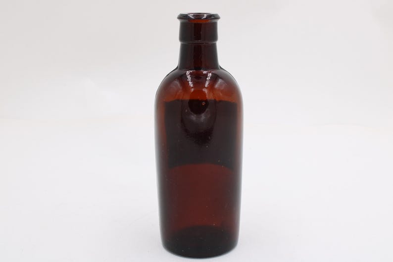 Puede incluir: Una botella de vidrio marr&oacute;n con un cuerpo redondeado y un cuello estrecho. La botella est&aacute; vac&iacute;a y tiene un color &aacute;mbar oscuro. La botella probablemente se utiliza para almacenar l&iacute;quidos.
