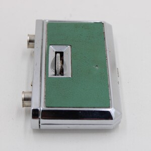 May include: A vintage, rectangular device with a chrome-colored metal frame and a textured, green center panel. It features a small dial and two silver-colored connectors. The device is likely an electronic component or accessory.