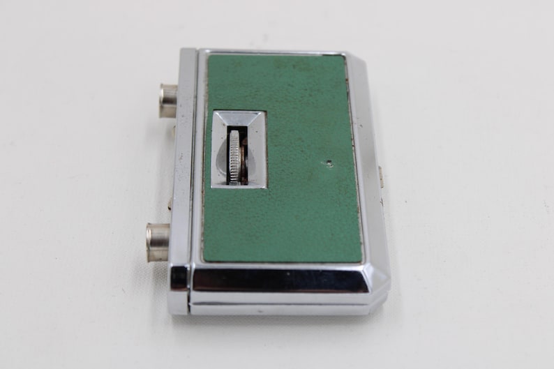 May include: A vintage, rectangular device with a chrome-colored metal frame and a textured, green center panel. It features a small dial and two silver-colored connectors. The device is likely an electronic component or accessory.