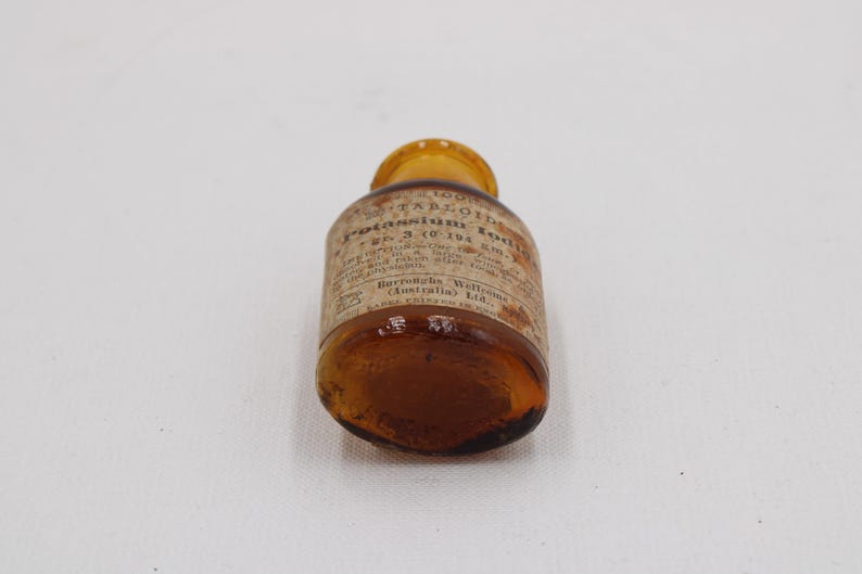 Puede incluir: Peque&ntilde;a botella de vidrio &aacute;mbar con tapa amarilla. La etiqueta, parcialmente visible, indica "Tabloids Potassium Iodide". La botella est&aacute; sobre una superficie blanca. Se aprecia el contenido.