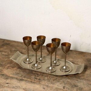 May include: Set of six silver shot glasses on a silver tray. The glasses have a gold interior and are arranged in a row on the tray.