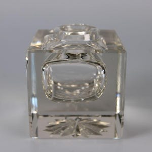 May include: A clear, rectangular crystal inkwell with a rounded front and a circular opening on top. The base features a decorative, etched floral design. The inkwell is made of transparent glass.