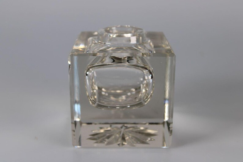 May include: A clear, rectangular crystal inkwell with a rounded front and a circular opening on top. The base features a decorative, etched floral design. The inkwell is made of transparent glass.