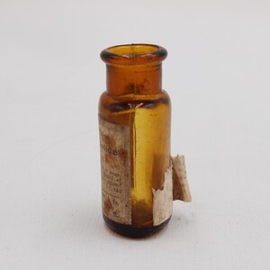 Puede incluir: Peque&ntilde;a botella de vidrio &aacute;mbar con base redondeada y cuello estrecho. La botella tiene una etiqueta con texto descolorido y un trozo de papel rasgado. Probablemente un art&iacute;culo antiguo o vintage.