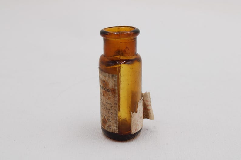 Puede incluir: Peque&ntilde;a botella de vidrio &aacute;mbar con base redondeada y cuello estrecho. La botella tiene una etiqueta con texto descolorido y un trozo de papel rasgado. Probablemente un art&iacute;culo antiguo o vintage.