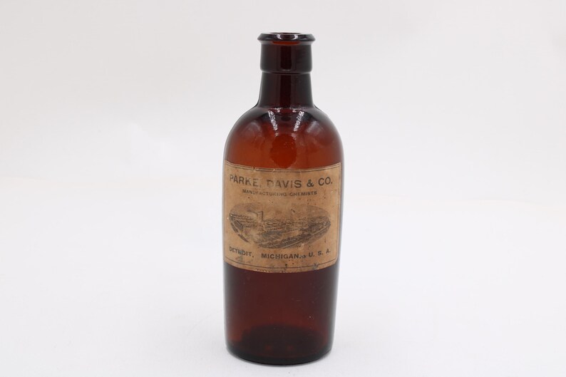 Puede incluir: Una botella de vidrio marr&oacute;n vintage con una etiqueta que dice "PARKE, DAVIS & CO. MANUFACTURING CHEMISTS DETROIT, MICHIGAN, U.S.A." La botella tiene un cuerpo redondeado y un cuello estrecho.