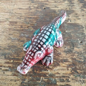 May include: A ceramic alligator figurine with a glossy finish. The alligator is painted with a red, white, and teal color scheme. The figurine is lying on a wooden surface.