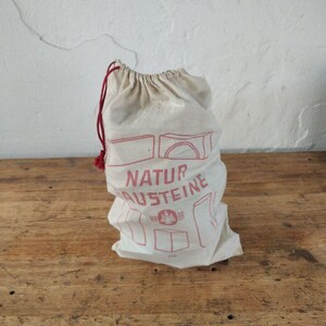 Könnte beinhalten: Ein beiger Kordelzugbeutel mit rotem Text und Illustrationen. Der Beutel ist gefüllt und steht auf einer Holzoberfläche. Der Text auf der Tasche lautet "NATUR AUSTEINE". Der Beutel ist mit einer roten Kordel gebunden.
