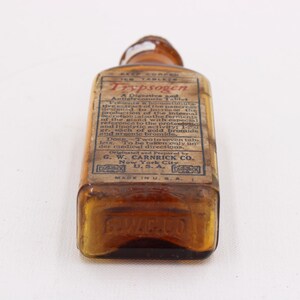 May include: An antique amber glass bottle with a rectangular label. The label reads "Trypsogen" and includes detailed text about the product. The bottle is marked "Made in U.S.A."