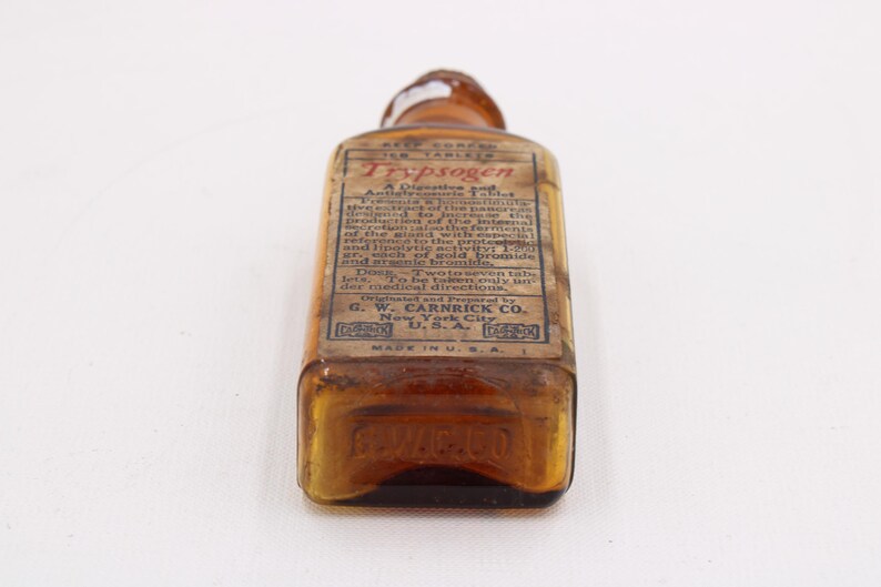 May include: An antique amber glass bottle with a rectangular label. The label reads "Trypsogen" and includes detailed text about the product. The bottle is marked "Made in U.S.A."