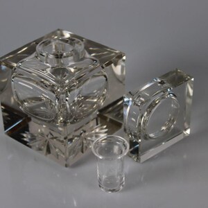 May include: Clear glass inkwell set. The set includes a large square inkwell with a circular opening, a smaller square piece, and a small cylindrical cup. The inkwell has a decorative, faceted design.