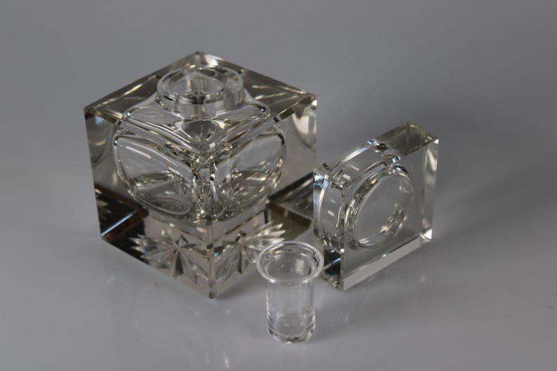 May include: Clear glass inkwell set. The set includes a large square inkwell with a circular opening, a smaller square piece, and a small cylindrical cup. The inkwell has a decorative, faceted design.