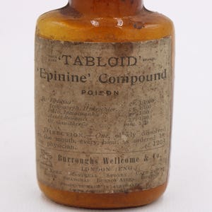 May include: An antique amber glass bottle with a paper label. The label reads "TABLOID Epinine Compound POISON" with detailed text and directions. The bottle has a vintage aesthetic, suggesting it is an old pharmaceutical item.