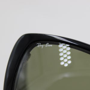 Puede incluir: Primer plano de gafas de sol Ray-Ban negras. El logotipo de Ray-Ban es visible en blanco en la montura. Las lentes son de color verde apagado. Las gafas de sol est&aacute;n sobre un fondo blanco liso.