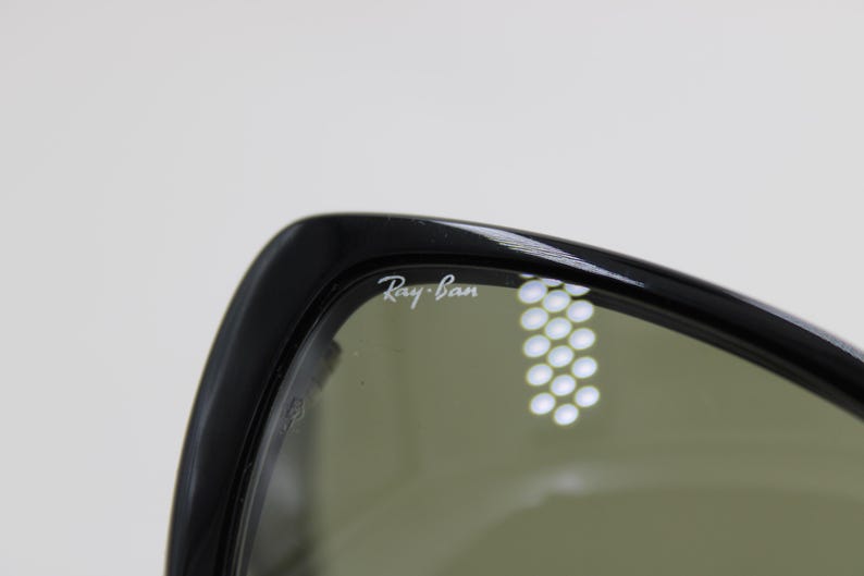 Puede incluir: Primer plano de gafas de sol Ray-Ban negras. El logotipo de Ray-Ban es visible en blanco en la montura. Las lentes son de color verde apagado. Las gafas de sol est&aacute;n sobre un fondo blanco liso.
