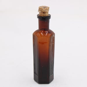 Puede incluir: Una botella de vidrio color &aacute;mbar con un tap&oacute;n de corcho. La botella tiene forma rectangular con detalles acanalados verticales. La botella es probablemente vintage y podr&iacute;a usarse con fines decorativos.