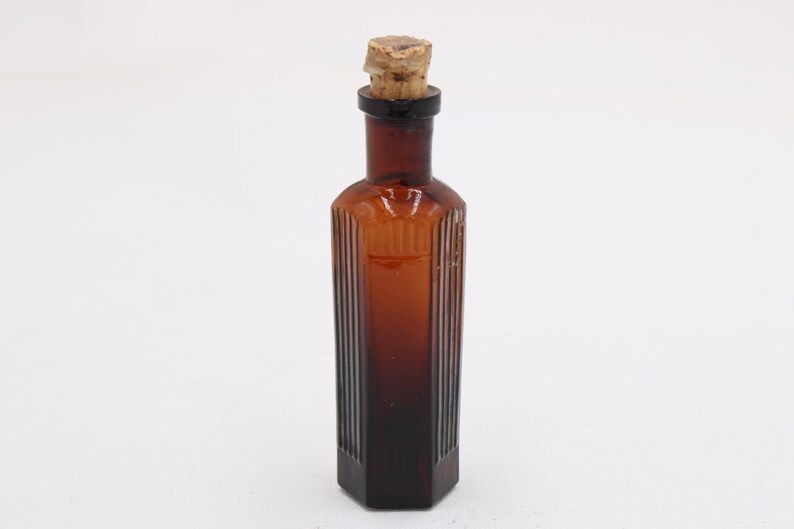 Puede incluir: Una botella de vidrio color &aacute;mbar con un tap&oacute;n de corcho. La botella tiene forma rectangular con detalles acanalados verticales. La botella es probablemente vintage y podr&iacute;a usarse con fines decorativos.