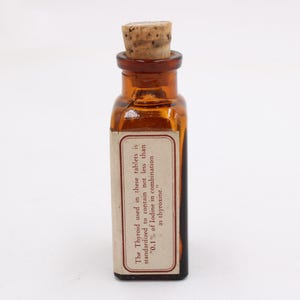 May include: A small, amber-colored glass bottle with a cork stopper. A rectangular label is affixed to the bottle, with text that reads: "The Thyroid used in these tablets is standardized to contain not less than '0.1% of Iodine in combination as thyroxine.'"