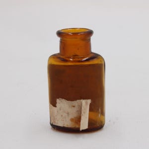 Puede incluir: Peque&ntilde;a botella de vidrio color &aacute;mbar, con cuello corto y cuerpo rectangular. Una etiqueta envejecida est&aacute; parcialmente adherida al frente. La botella est&aacute; vac&iacute;a y se encuentra sobre un fondo blanco.