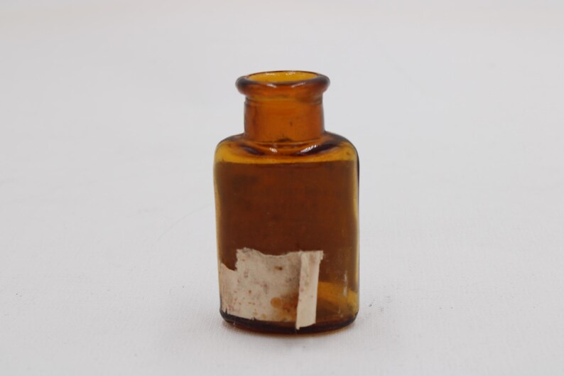 Puede incluir: Peque&ntilde;a botella de vidrio color &aacute;mbar, con cuello corto y cuerpo rectangular. Una etiqueta envejecida est&aacute; parcialmente adherida al frente. La botella est&aacute; vac&iacute;a y se encuentra sobre un fondo blanco.