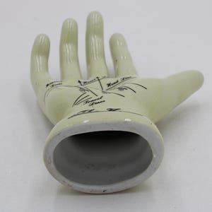 May include: Off-white ceramic hand figurine with black palmistry markings. The open hand shows lines labelled "Heart Lines", "Travel Lines", and "Head Lines". The base is a hollow oval shape.