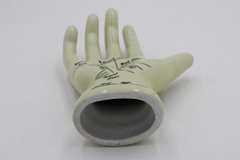 May include: Off-white ceramic hand figurine with black palmistry markings. The open hand shows lines labelled "Heart Lines", "Travel Lines", and "Head Lines". The base is a hollow oval shape.