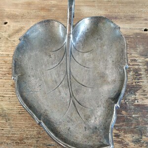 Peut inclure: Un plat argent&eacute; en forme de feuille avec une poign&eacute;e. Le plat pr&eacute;sente un motif de veines d&eacute;taill&eacute; et un aspect l&eacute;g&egrave;rement terni, sugg&eacute;rant l'&acirc;ge. La poign&eacute;e est incurv&eacute;e et fix&eacute;e au centre de la feuille.