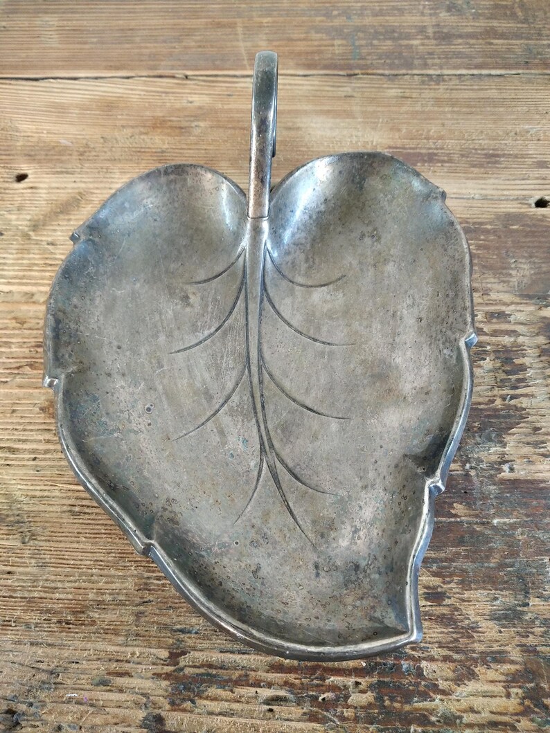 Peut inclure: Un plat argent&eacute; en forme de feuille avec une poign&eacute;e. Le plat pr&eacute;sente un motif de veines d&eacute;taill&eacute; et un aspect l&eacute;g&egrave;rement terni, sugg&eacute;rant l'&acirc;ge. La poign&eacute;e est incurv&eacute;e et fix&eacute;e au centre de la feuille.