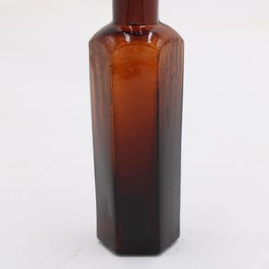 Puede incluir: Una botella de vidrio marr&oacute;n vintage con una base hexagonal y un cuerpo largo y delgado. La botella tiene un color &aacute;mbar oscuro que se desvanece a un tono m&aacute;s claro hacia la parte superior. La botella est&aacute; vac&iacute;a y tiene un acabado liso y brillante.