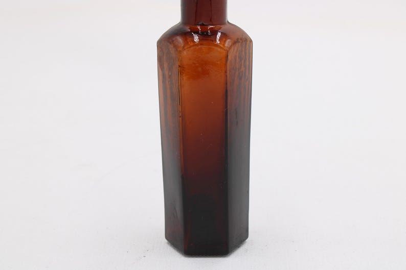 Puede incluir: Una botella de vidrio marr&oacute;n vintage con una base hexagonal y un cuerpo largo y delgado. La botella tiene un color &aacute;mbar oscuro que se desvanece a un tono m&aacute;s claro hacia la parte superior. La botella est&aacute; vac&iacute;a y tiene un acabado liso y brillante.